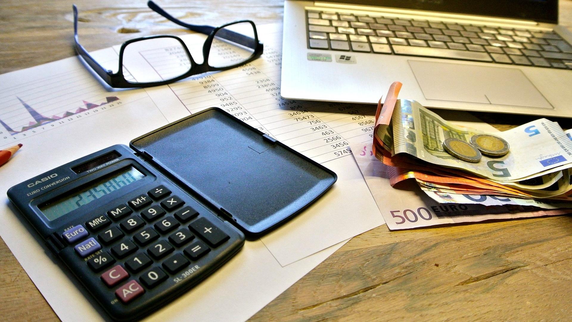 Blick auf einen Schreibtisch mit Laptop, Taschenrechner, Statistiken und Geld