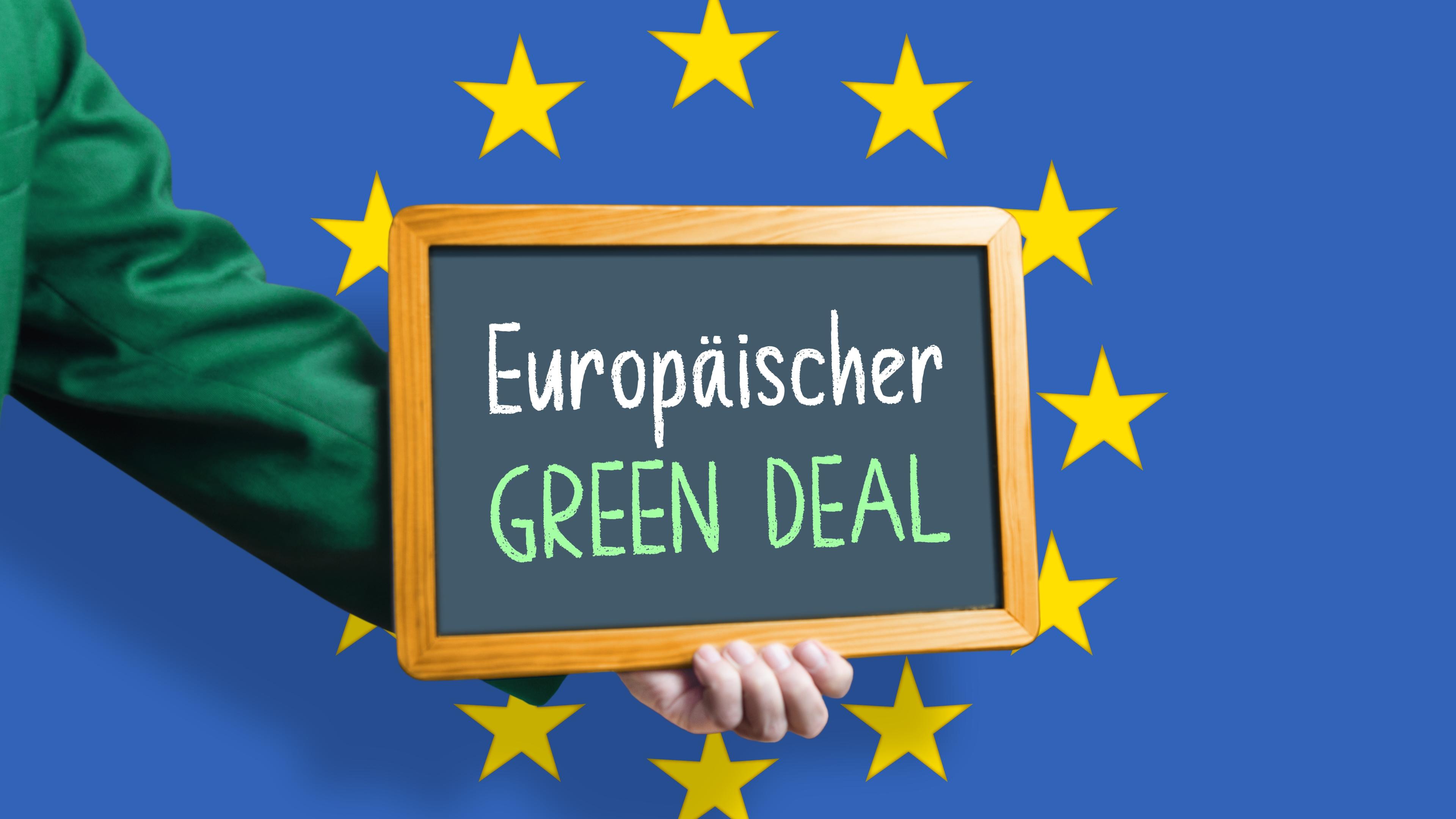 Ein grün bekleideter Arm hält eine Tafel mit Aufschrift "Europäischer Green Deal!" in den EU-Sternenkreis vor blaubem Hintergrund
