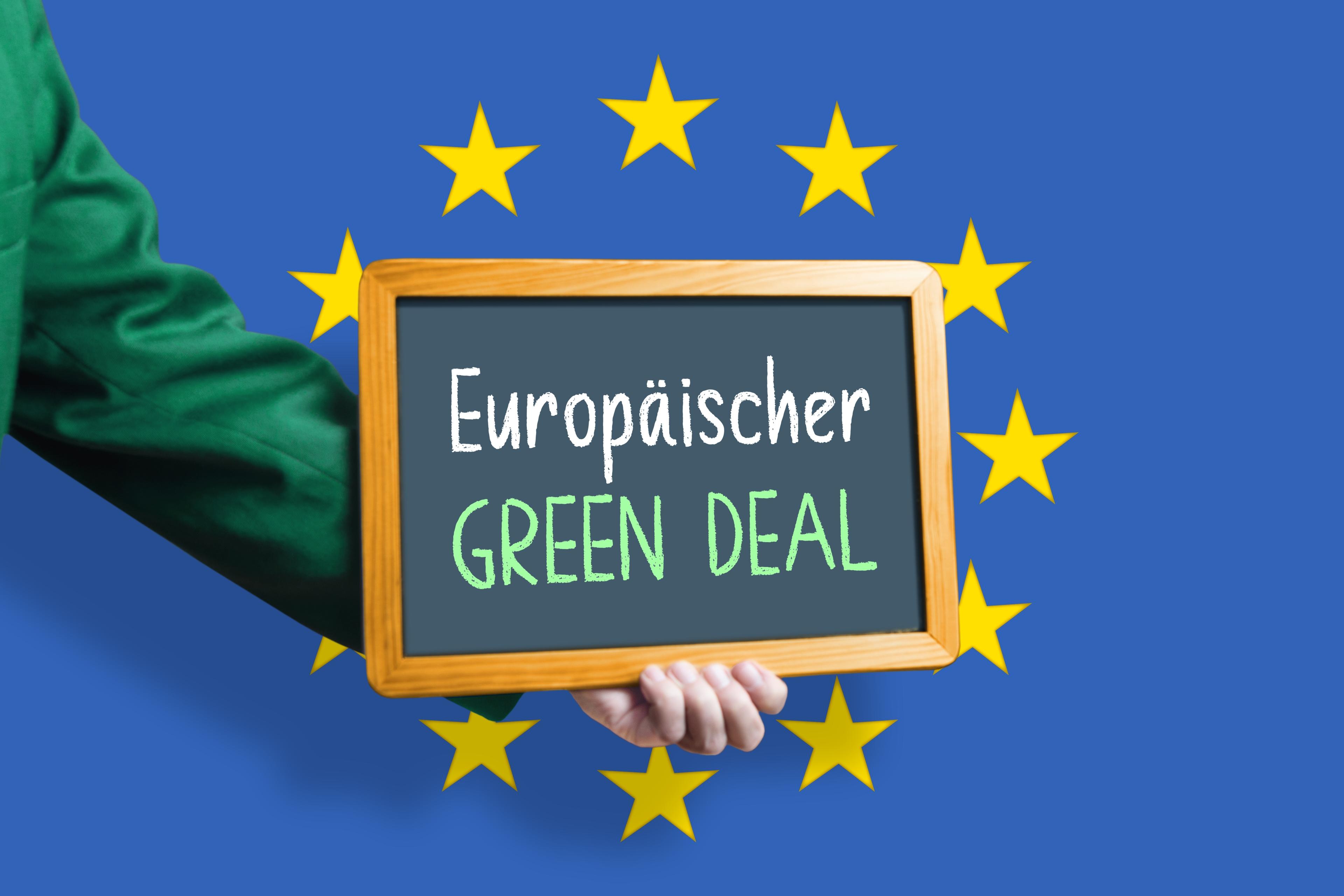 Ein grün bekleideter Arm hält eine Tafel mit Aufschrift "Europäischer Green Deal!" in den EU-Sternenkreis vor blaubem Hintergrund